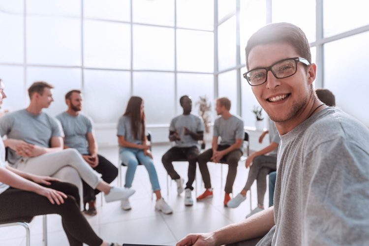 smiling young man sitting in circle with group - group therapy