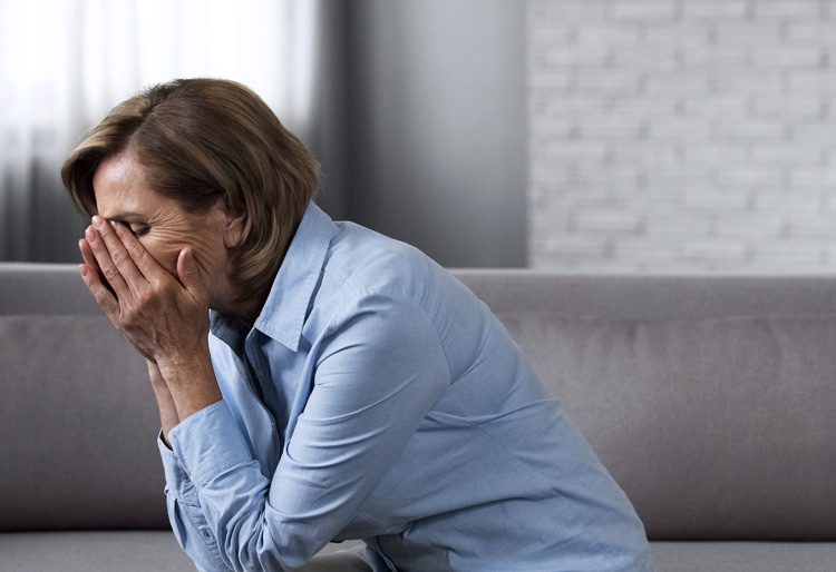 senior woman sitting at home on the edge of the couch with her face in her hands - anxiety