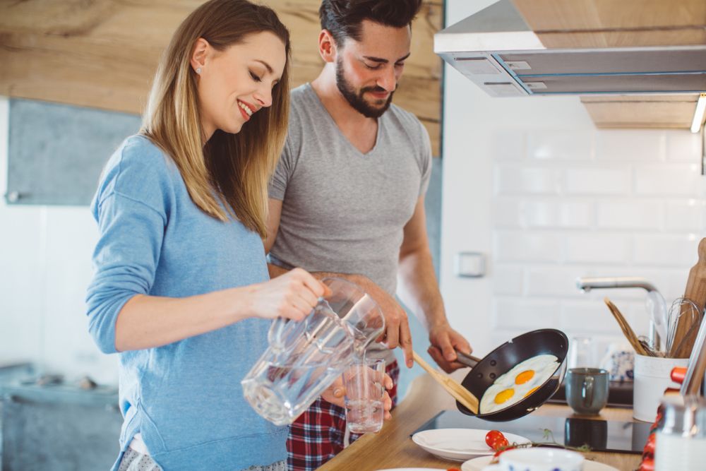Smiling couple making breakfast together—man cooking eggs while woman pours water into a glass in a bright kitchen.