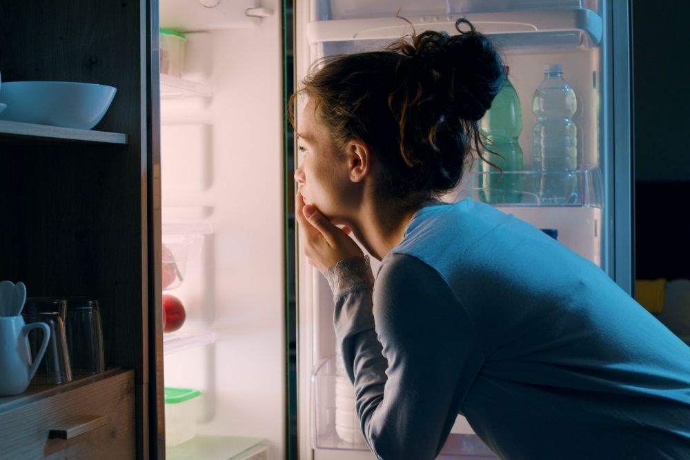 A young woman in pajamas looks thoughtfully into an open refrigerator at night, deciding what to eat