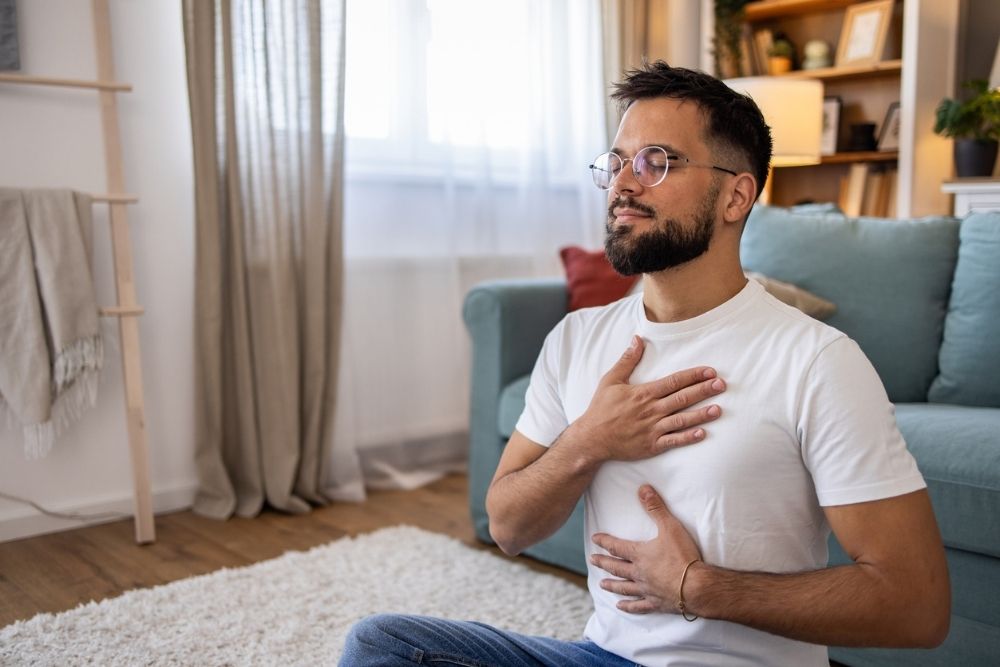 Man sitting cross-legged on the floor with eyes closed and hands on chest and abdomen, practicing mindful breathing in a peaceful home environment.