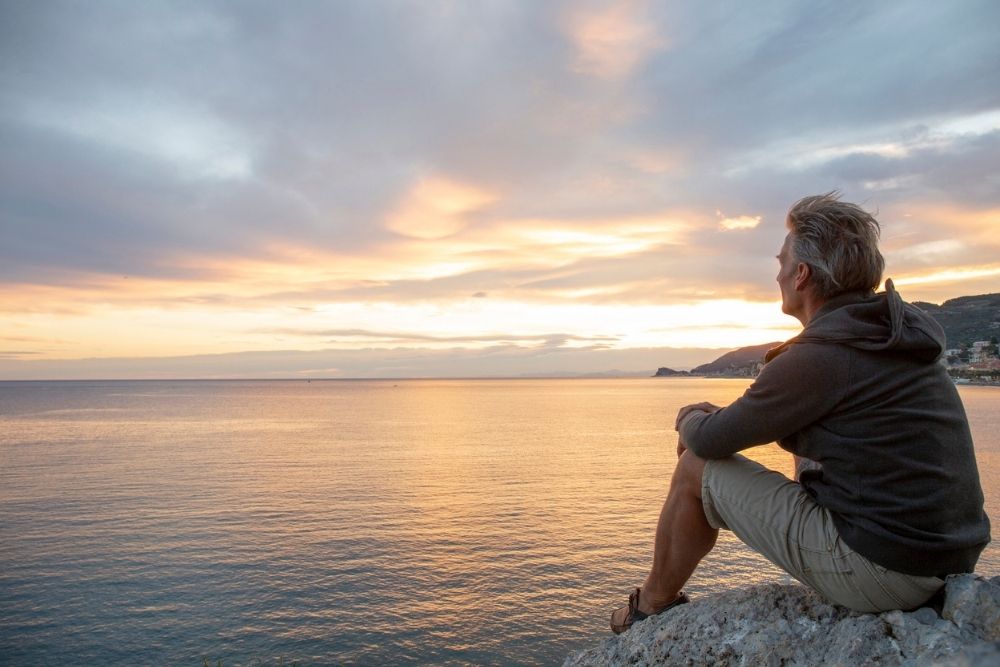 Man sitting on a rock by the sea at sunset, looking out thoughtfully over the water.