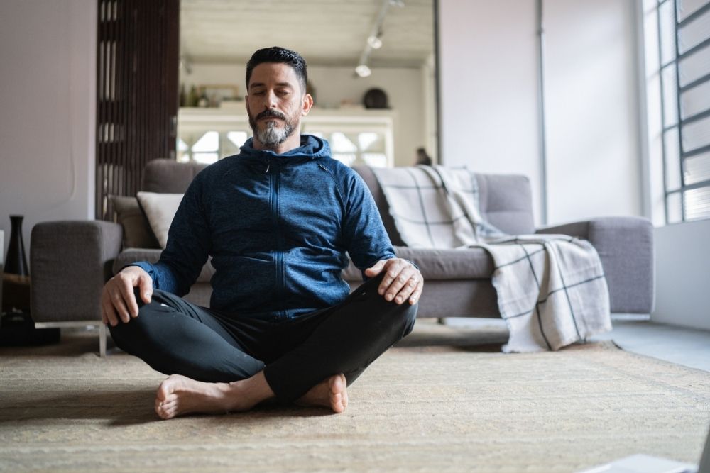 Man sitting cross-legged on the floor at home with eyes closed, practicing mindfulness or meditation for relaxation and balance.
