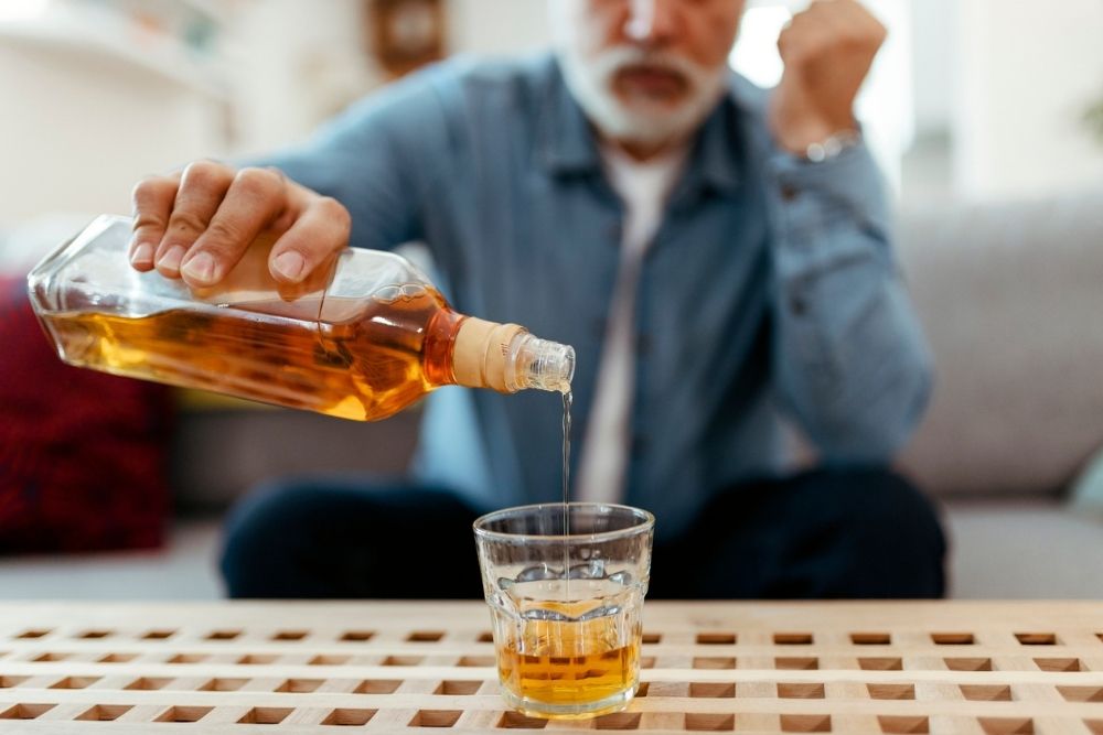 A person pours liquor from a bottle into a glass while sitting on a couch, with their face slightly out of focus in the background.