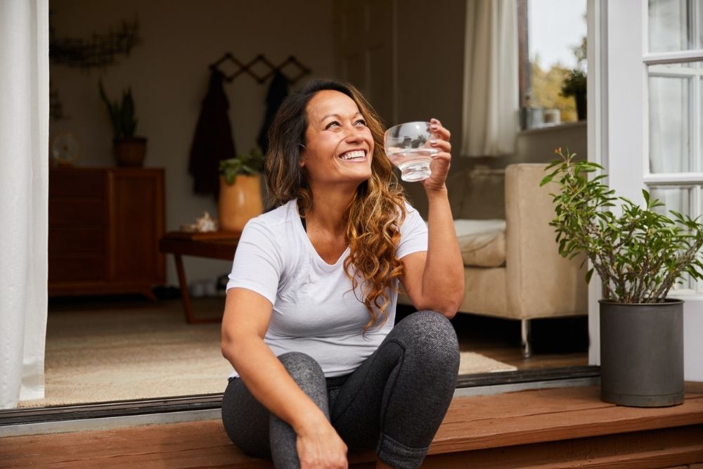 A smiling woman sits on a doorstep holding a glass of water, looking upward. She appears relaxed and content outside a cozy home interior.