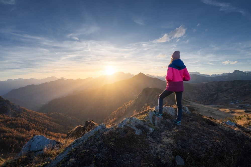 A person standing on a mountain ridge at sunrise, looking out over a vast landscape of peaks and valleys.